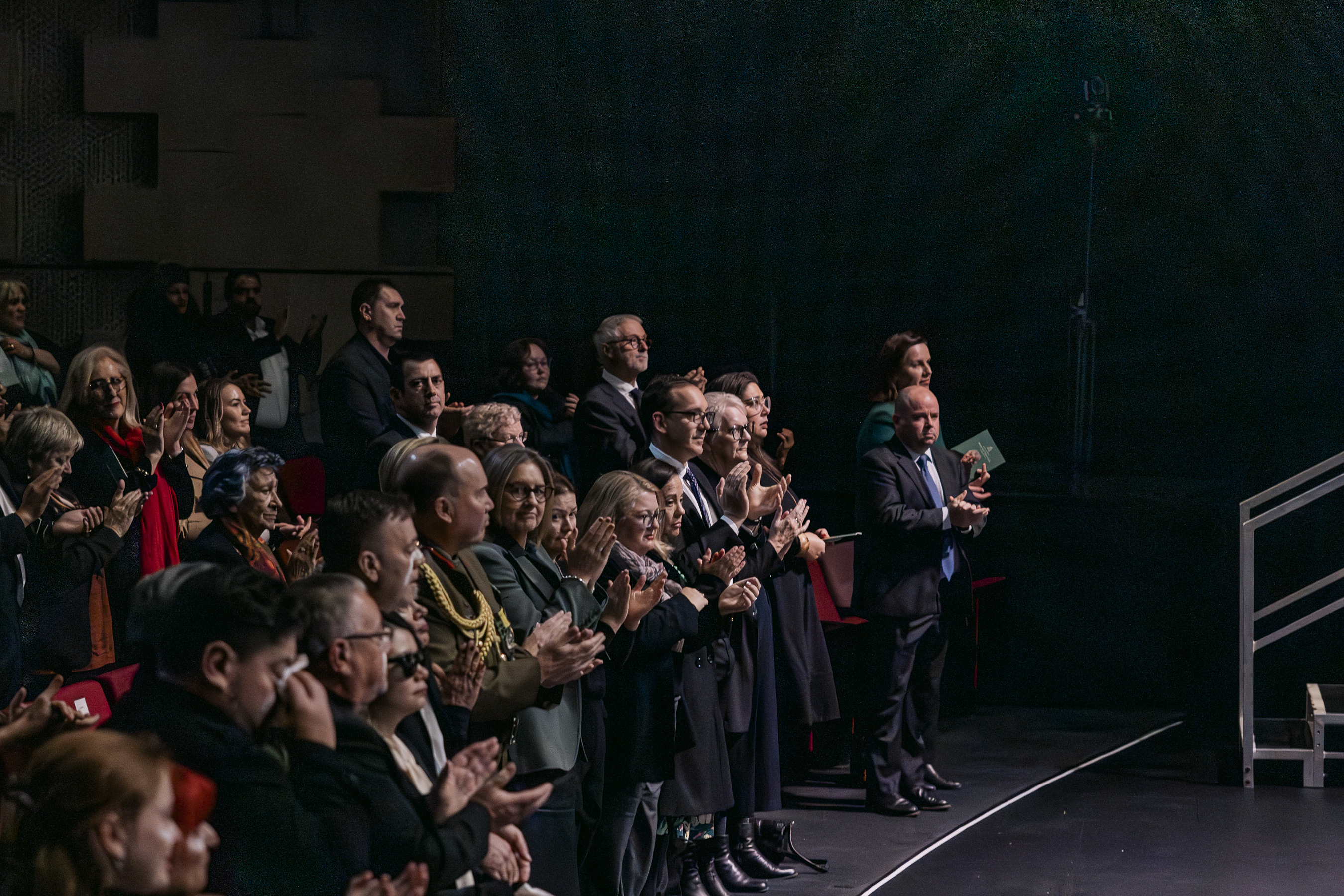 People in the audience of a dark theatre stand and clap. 