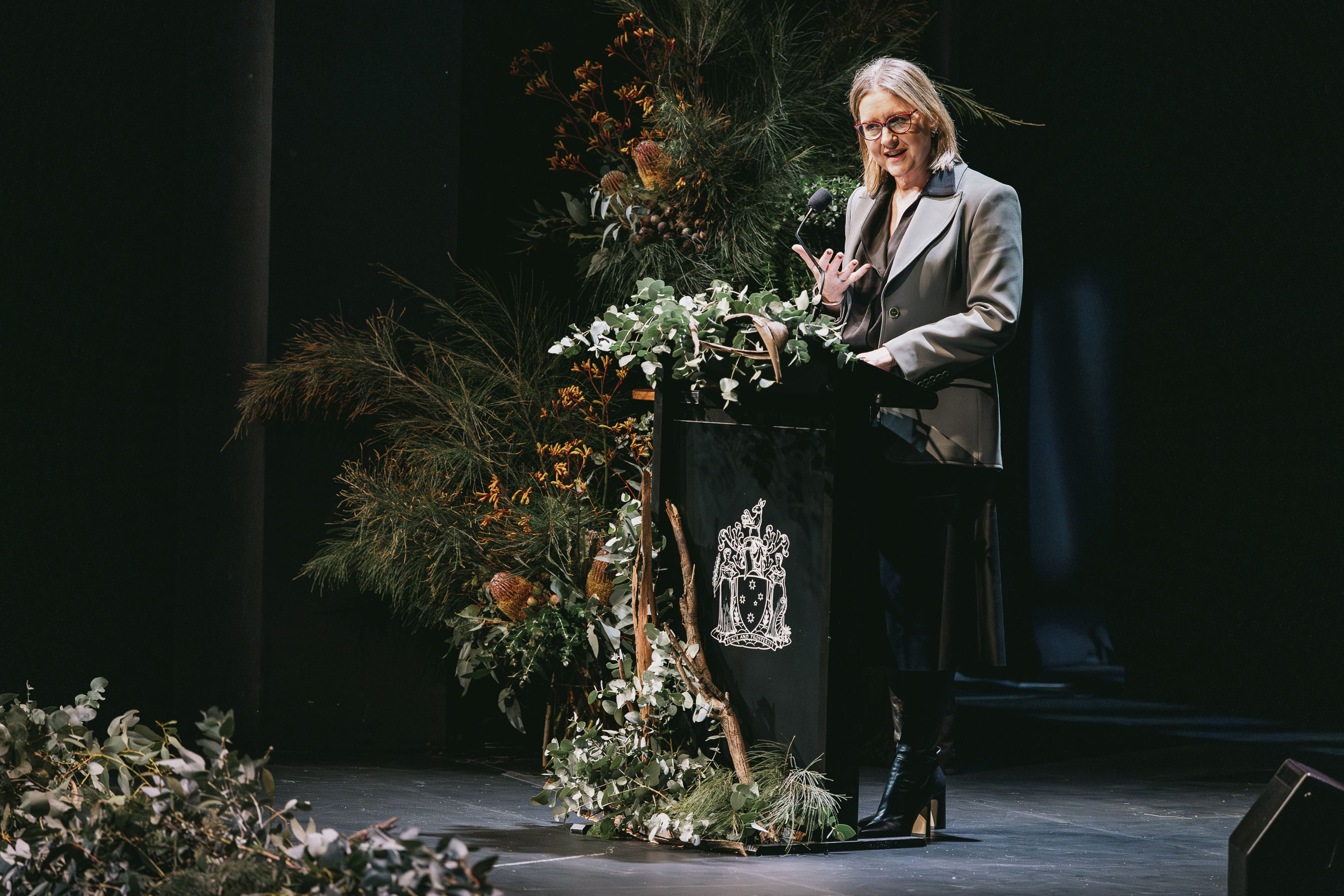 A person in a grey suit jacket and blonde hair speaks at a podium that is covered in flowers. 