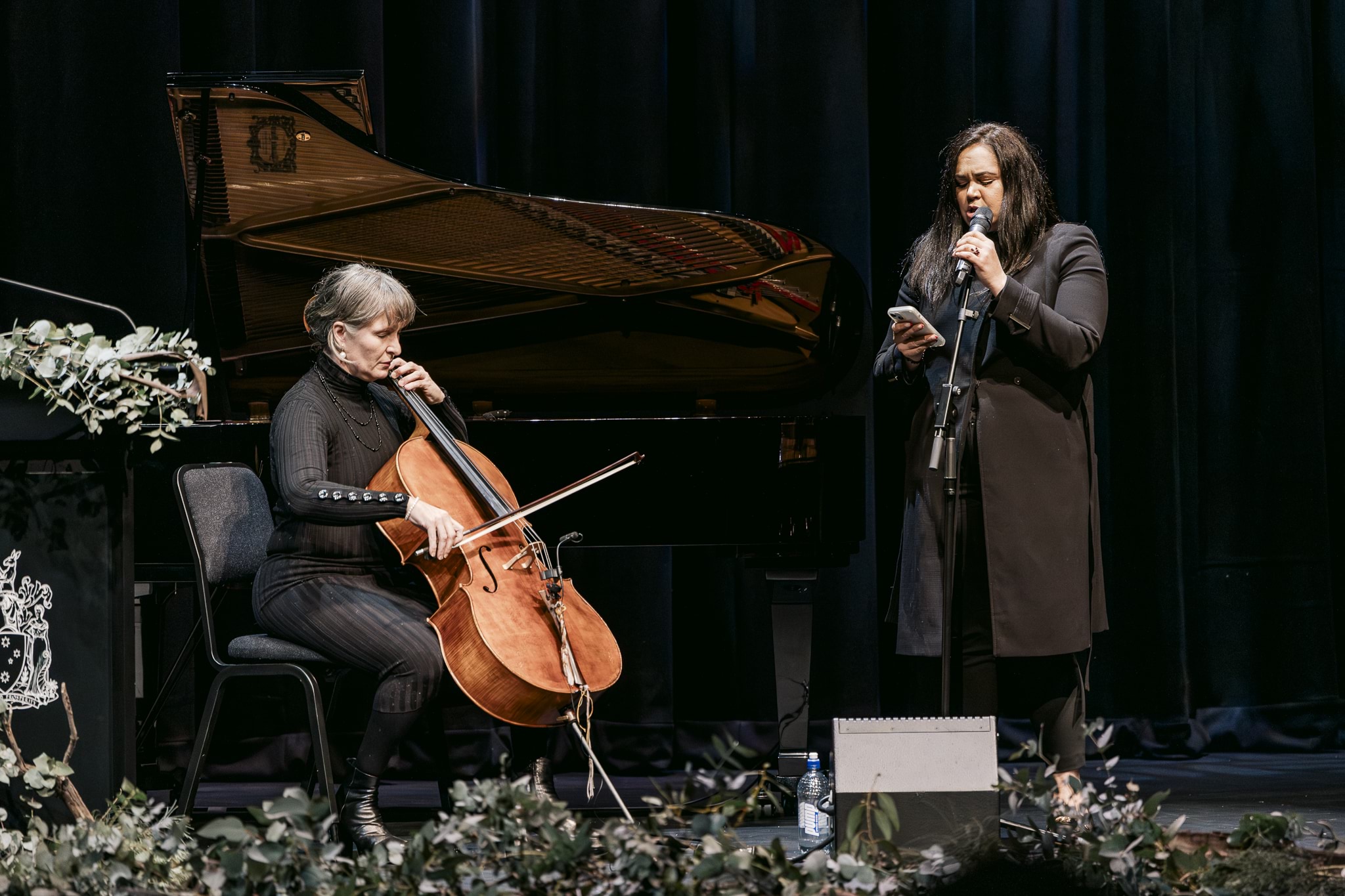 A person playing a cello and a person at a microphone on a stage with flowers and a piano. They are both wearing black. 