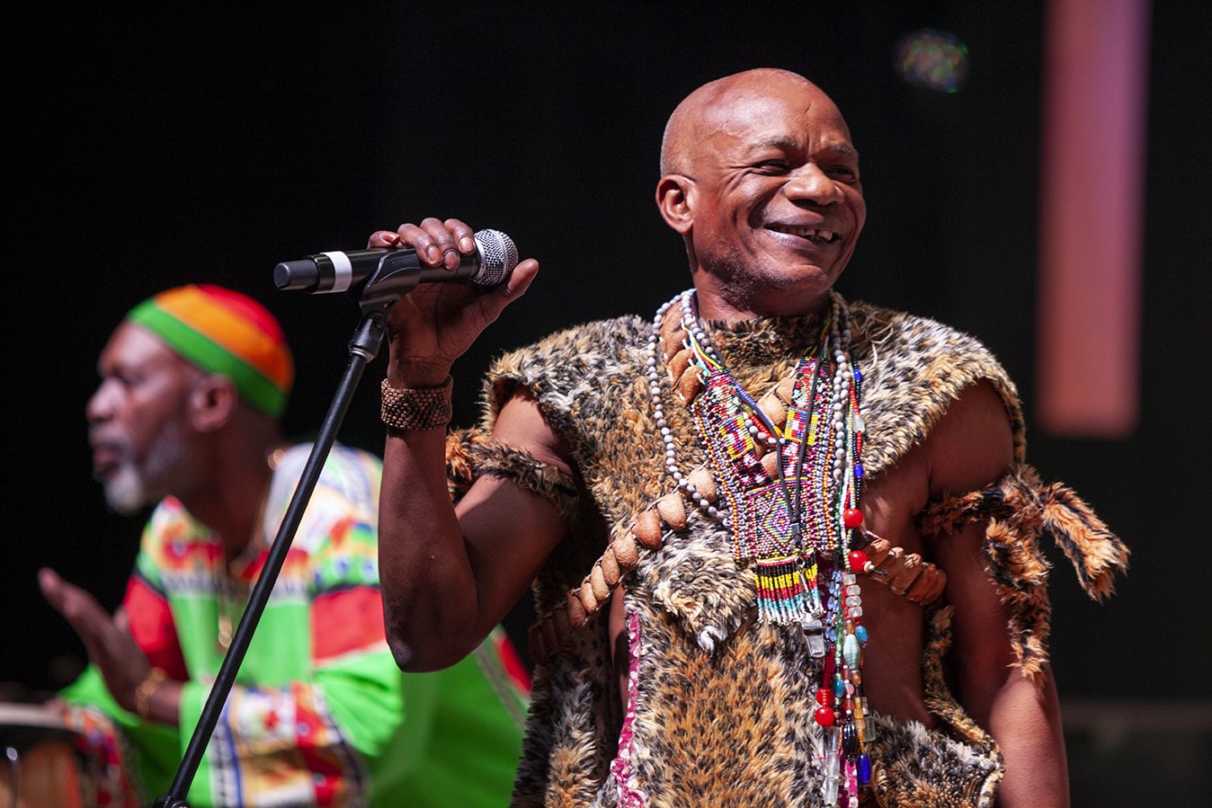 A male musician of African background wearing colourful costume smiles in front of a microphone, while another plays drums in the background