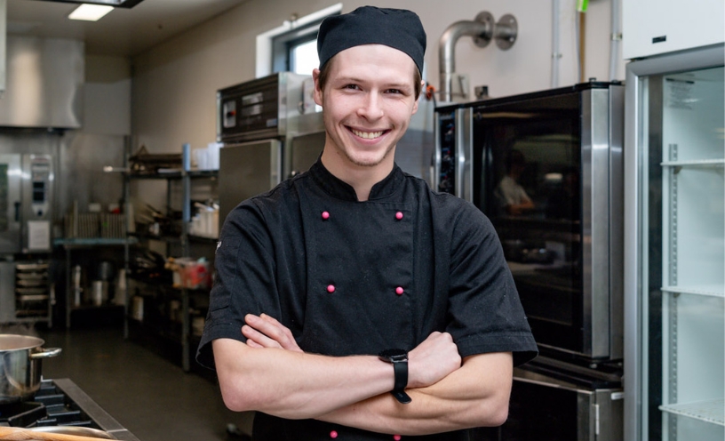 A chef in a black jacket and hat smiling with arms crossed in a commercial kitchen.