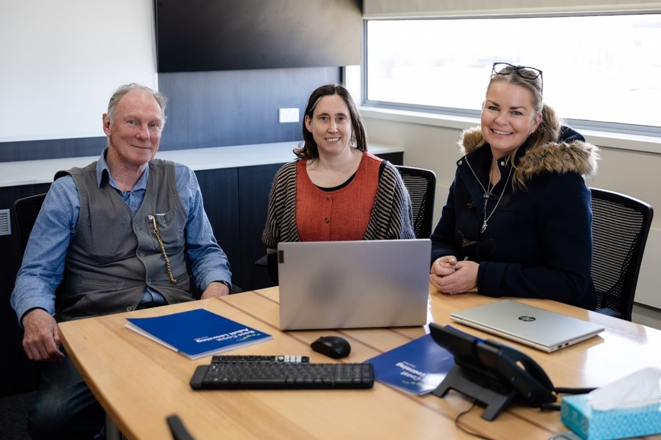 3 people sitting at a table, facing the camera, smiling, with a lap top in front of them