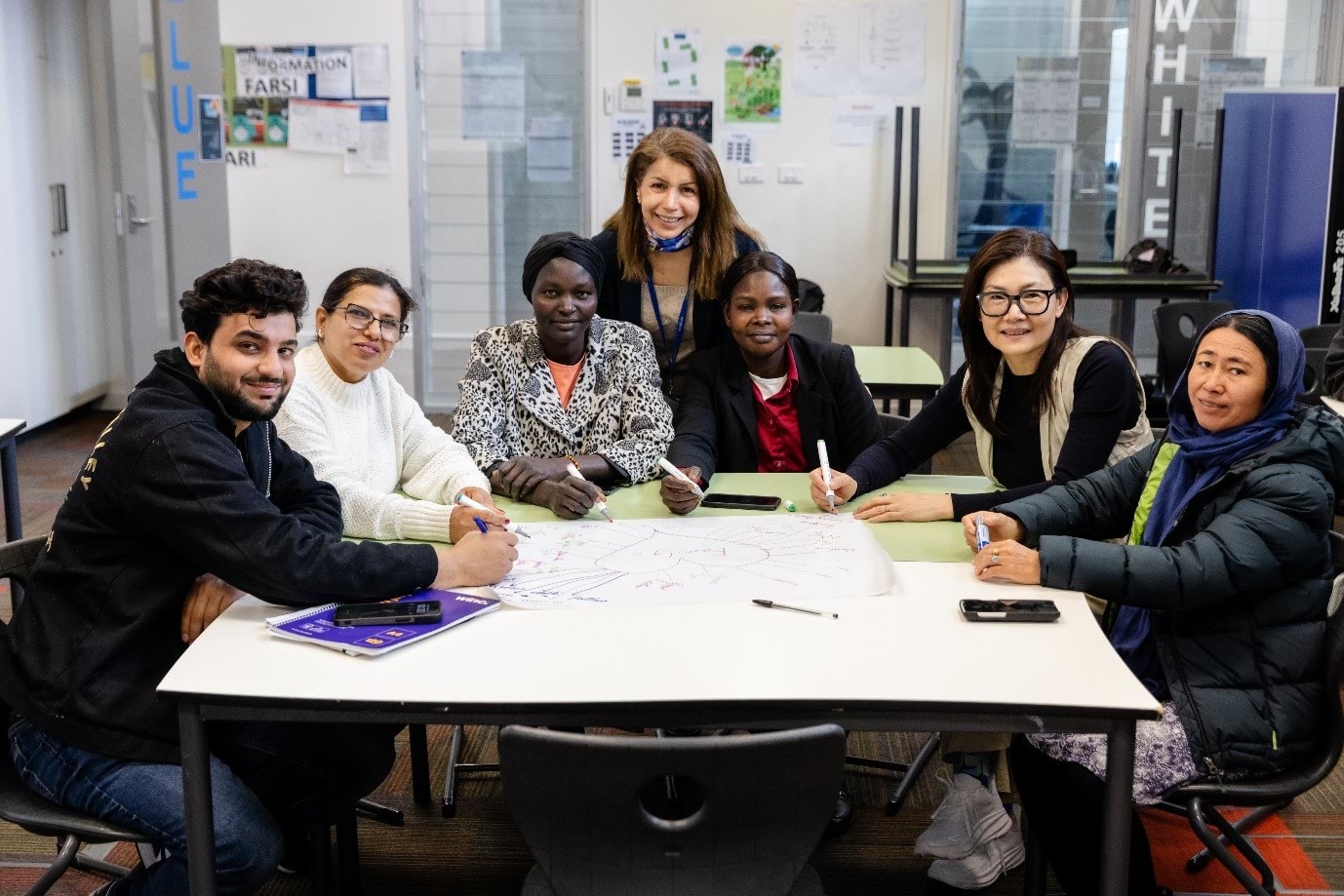 7 people sitting at a table, facing the camera and smiling, holding pens with a shared piece of paper.