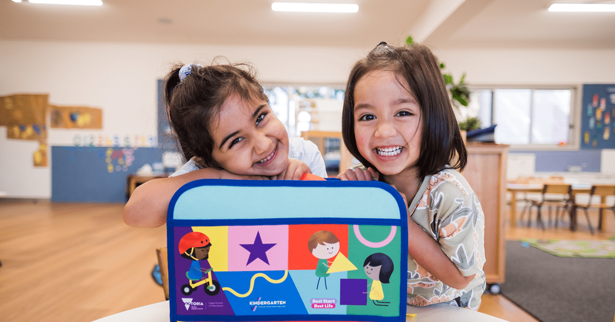 2 young children hold a colourful kinder kit in a kindergarten classroom.