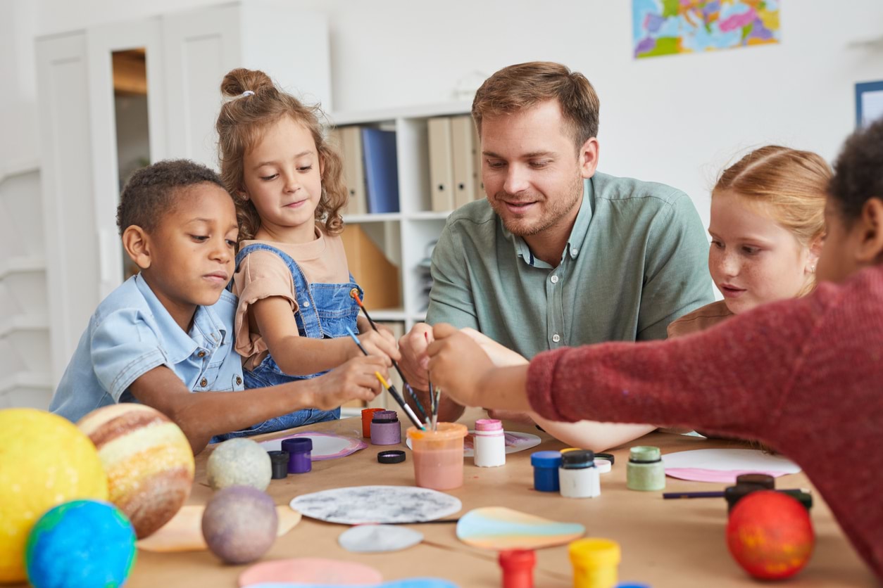 Children sitting at a table painting with their teacher.