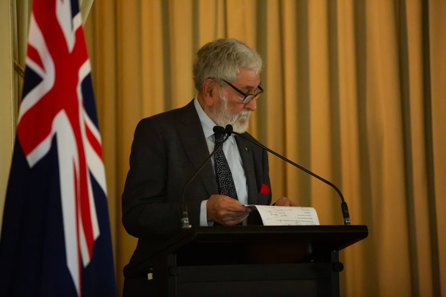 Hon. Dr Barry Jones stands at lectern reading from his notes, the Australian flag is behind him