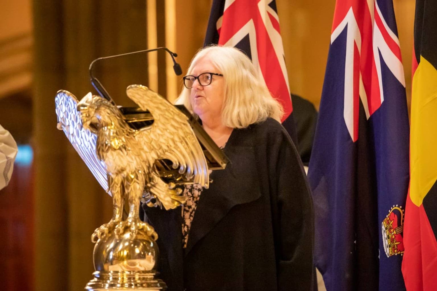 Aunty Di Kerr OAM speaks at a podium shaped like an eagle. There are flags behind her.
