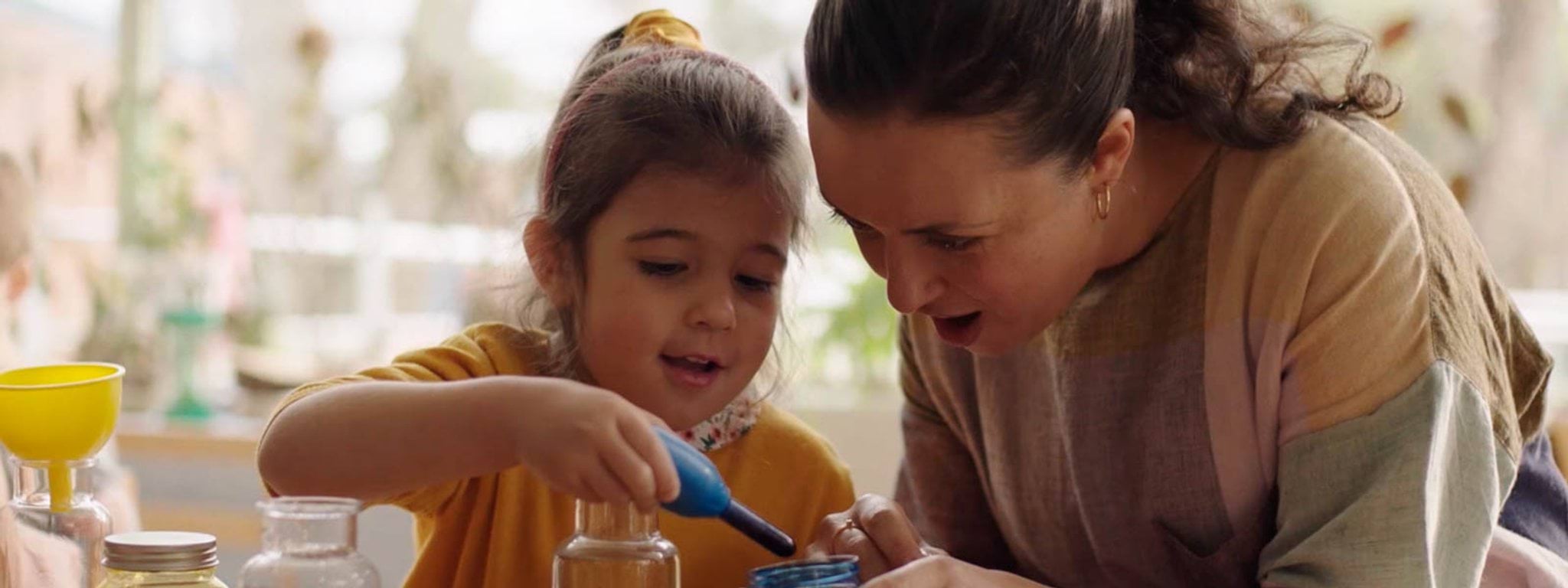 A woman helps a young child play with colourful dyes in a bright, sunny kindergarten
