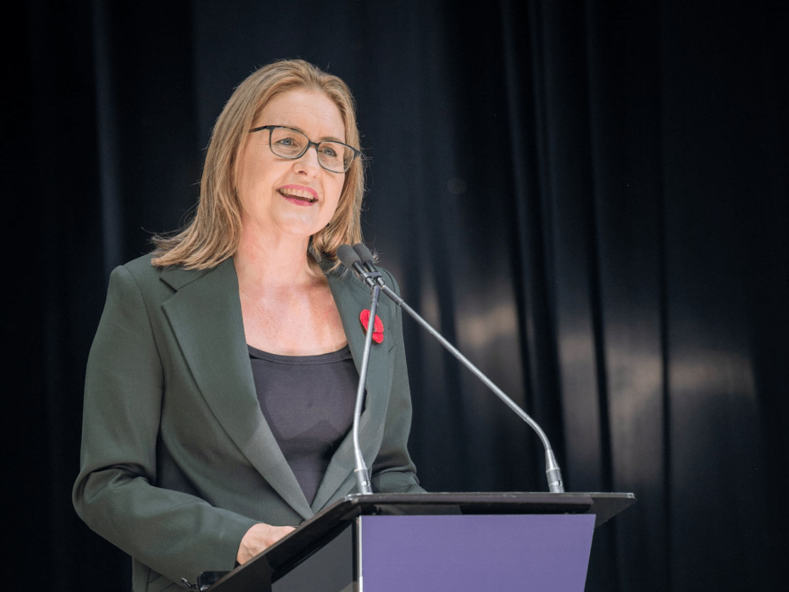 a woman in a dark green suit stands at a podium speaking to an audience