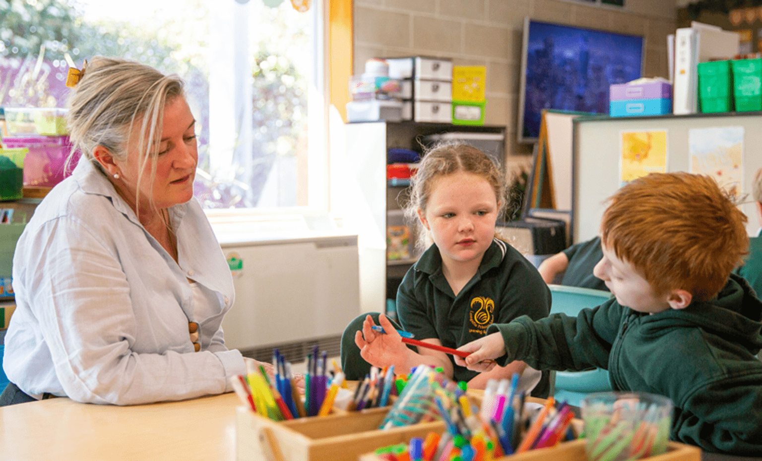 A female teacher talks to two children in green uniforms over a table with lots of colourful pens and pencils.