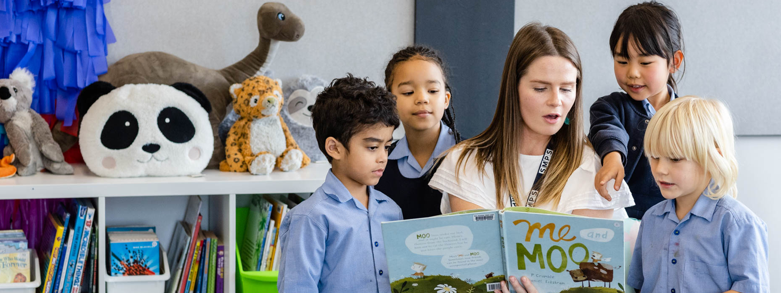 A teacher reads from a children's book with children gathered around her and surrounded by books and soft toys.