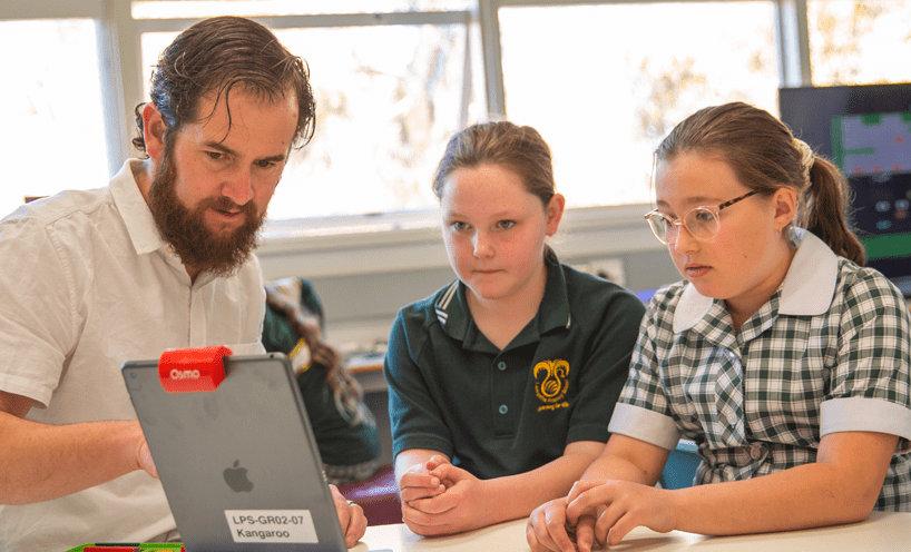 A male school teacher in a white shirt sits with two female students in a green t-shirt and a green and white school dress. They are all looking at an iPad and mid conversation.