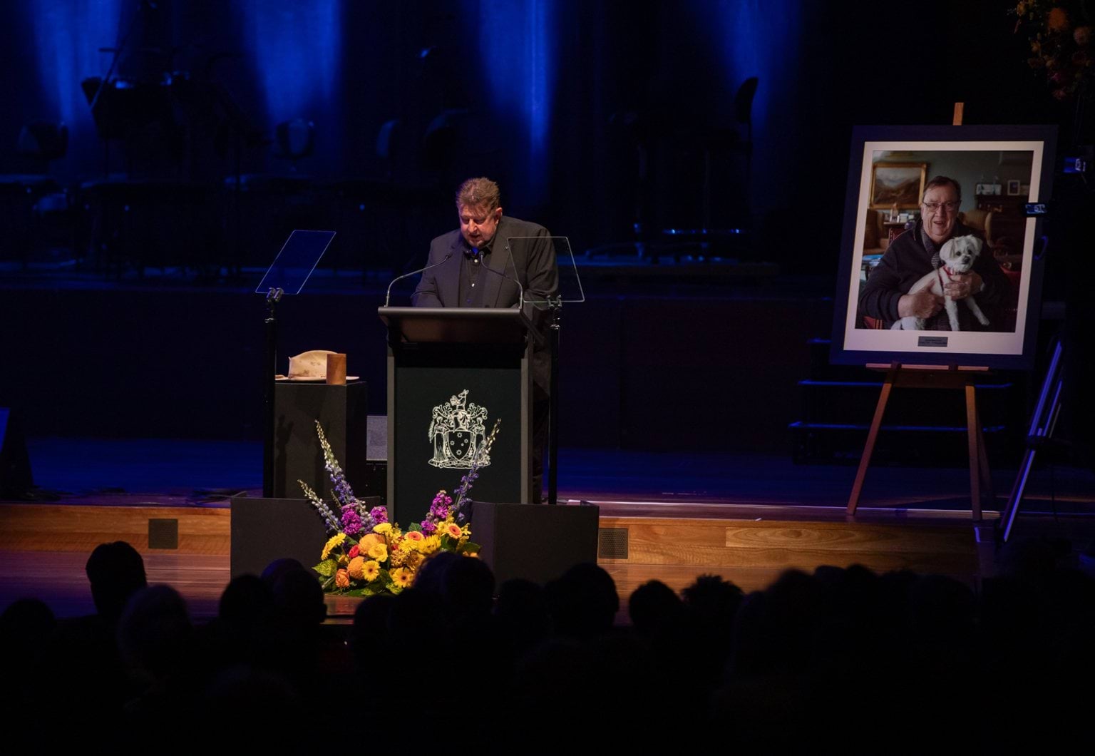 A man wearing black stands at a lectern on a stage with flowers and a framed photo of a man with a dog 