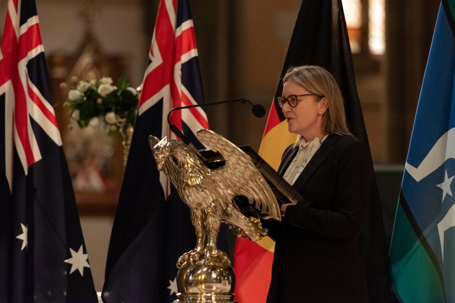 A woman with dark blonde hair in a white shirt and black blazer speaks into a microphone at a church pulpit, with the Australian, Victorian, Aboriginal and Torres Strait Islander flags in the background. 