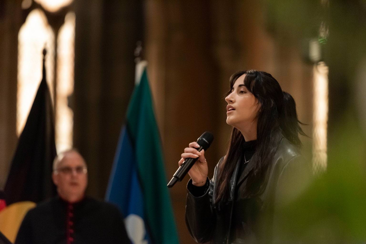 A woman with long black hair in all black clothing sings into a microphone.  