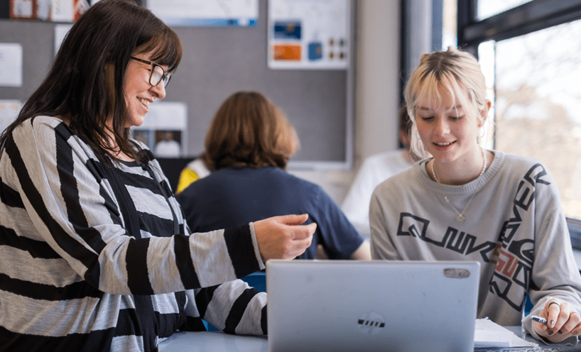 A female teacher in a grey and black striped shirt sits with a female high school student in a grey shirt. They are smiling down at a computer screen in front of them. 