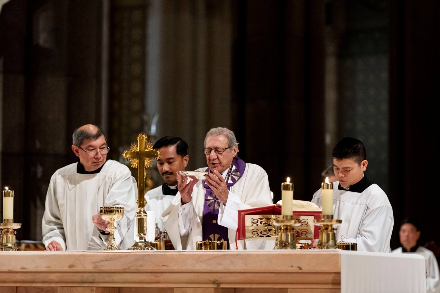 Four men wear white biblical dress and stand in front of an alter with a man positioned in the middle holding a silver communion plate.   