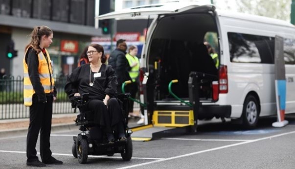 Photo of a person in a motorised wheelchair and a customer officer chat outside a wheelchair accessible taxi with the wheelchair lift down on the ground.