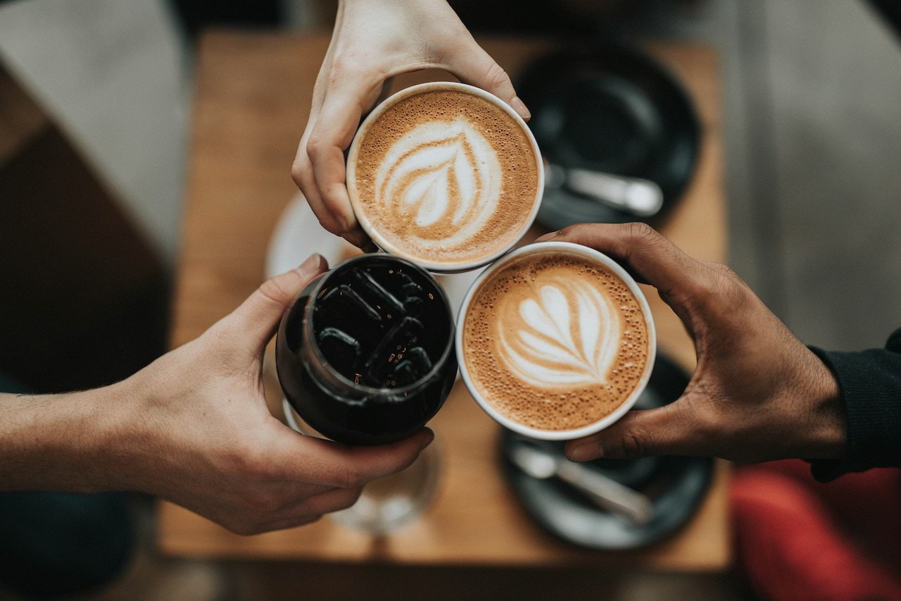 Two caffe lattes and a coke being clinked together in a cafe