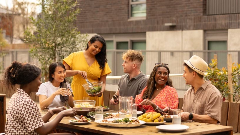 A group of 6 young people enjoy a meal on a shared balcony in a new apartment block