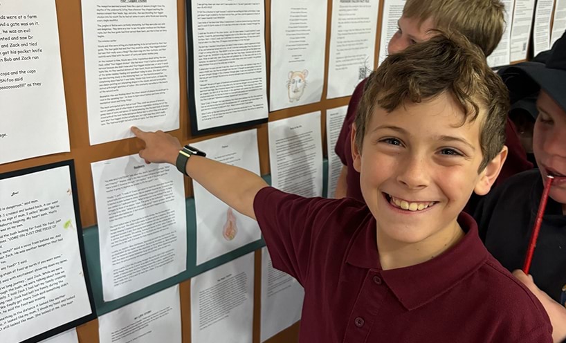 A smiling boy in a burgundy shirt points at a wall displaying various printed pages.