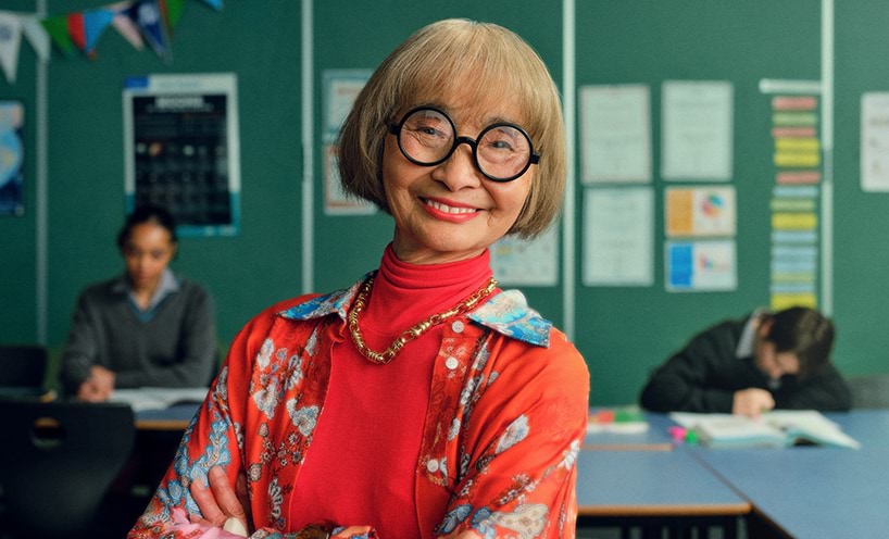 A teacher standing in a classroom wearing colourful clothes and round glasses.