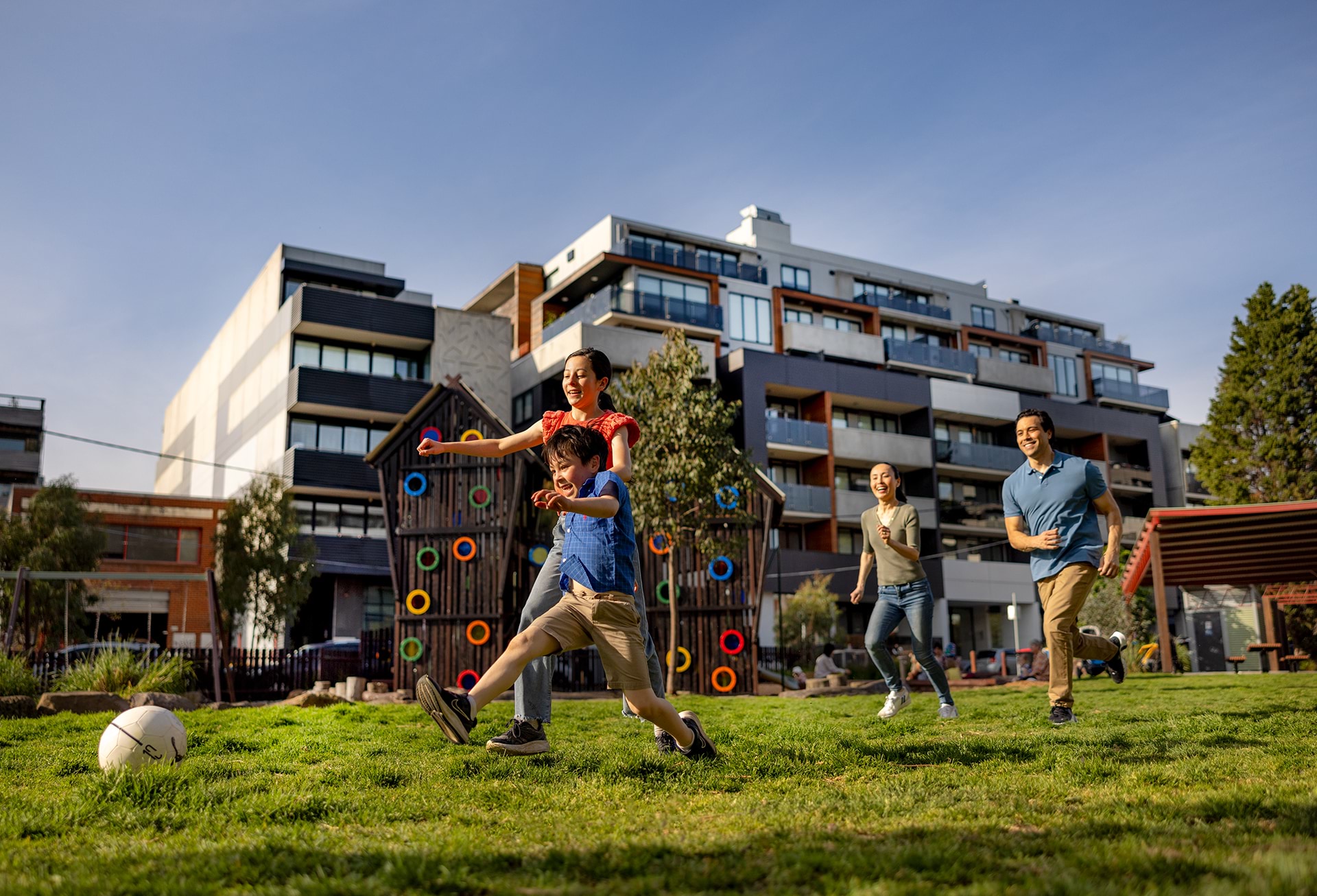 A smiling family of four plays soccer in a park with a 6-storey apartment block in the background.