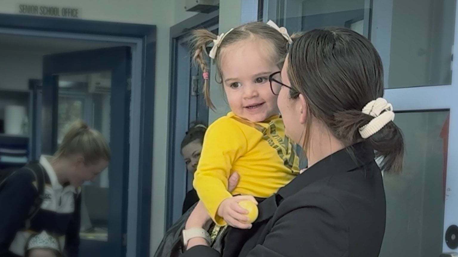 A woman holds a little girl wearing a yellow t-shirt, holding a yellow ball.