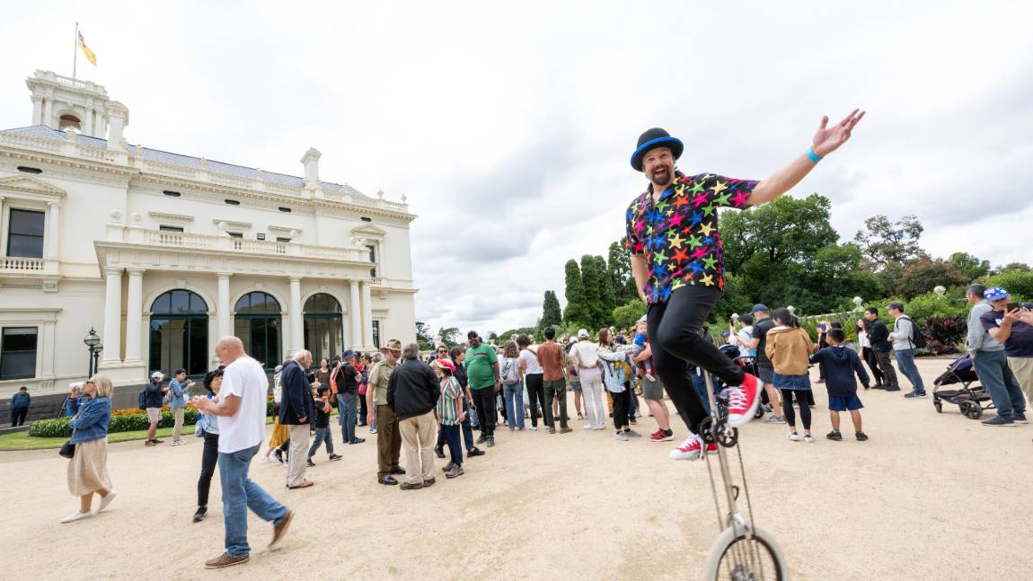 A smiling man wearing a shirt with multicoloured stars rides a unicycle on the grounds of Government House