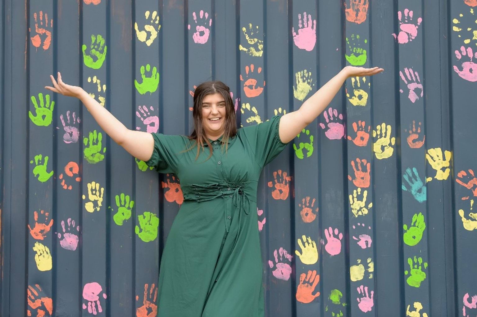 Pre-service teacher placement grants program A woman in a green dress stands with arms raised in front of a wall covered with colourful handprints.