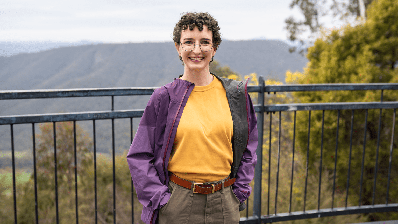 A woman in a yellow t-shirt and purple jacket stands on a bridge in front of mountains and trees.