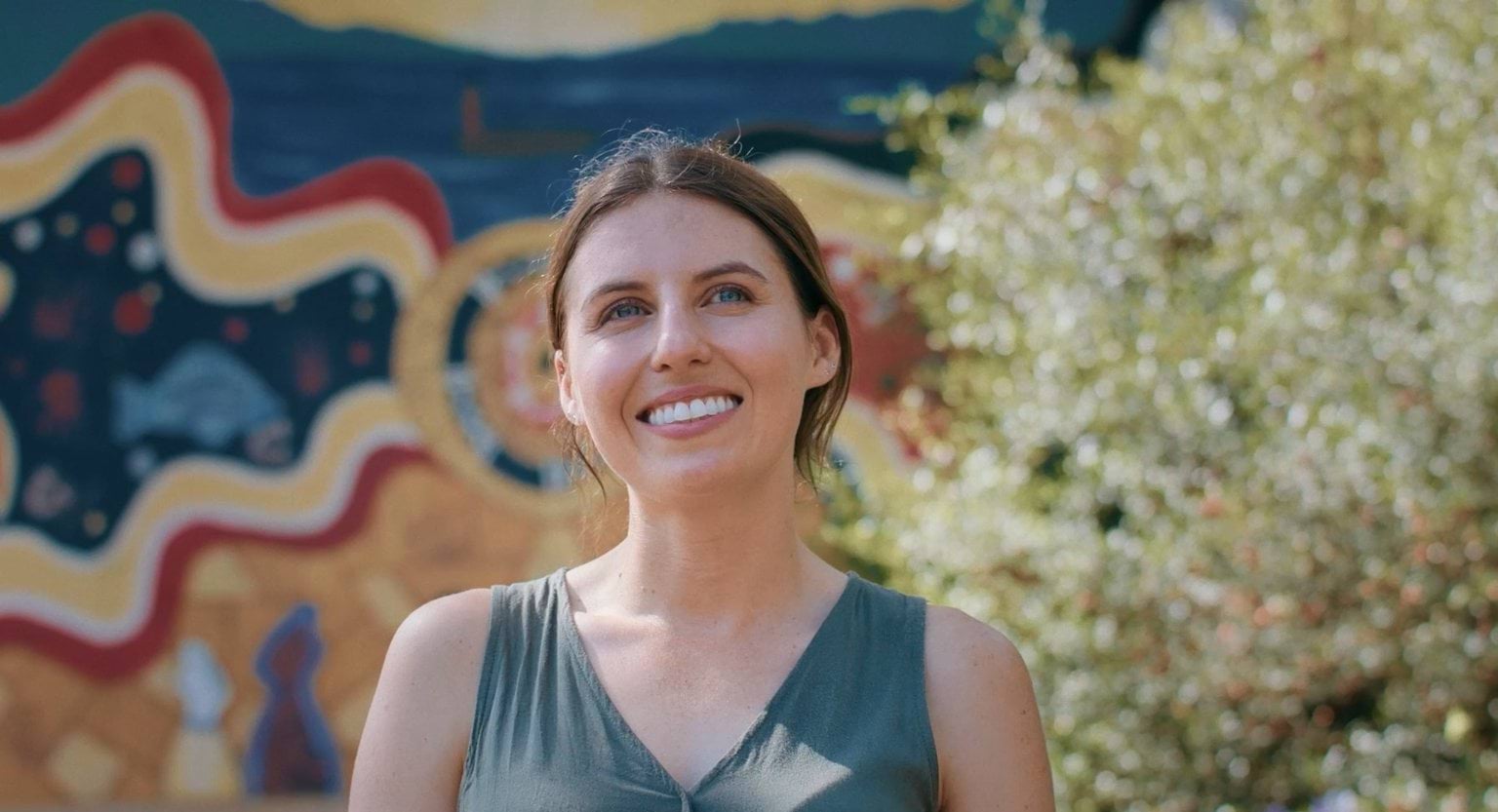 A woman wearing a green top stands in front of a colourful painted wall and a tree.