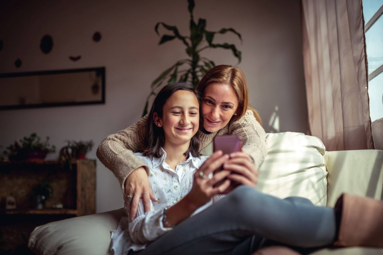 A woman and teenage girl sitting on the couch together, looking at a phone.