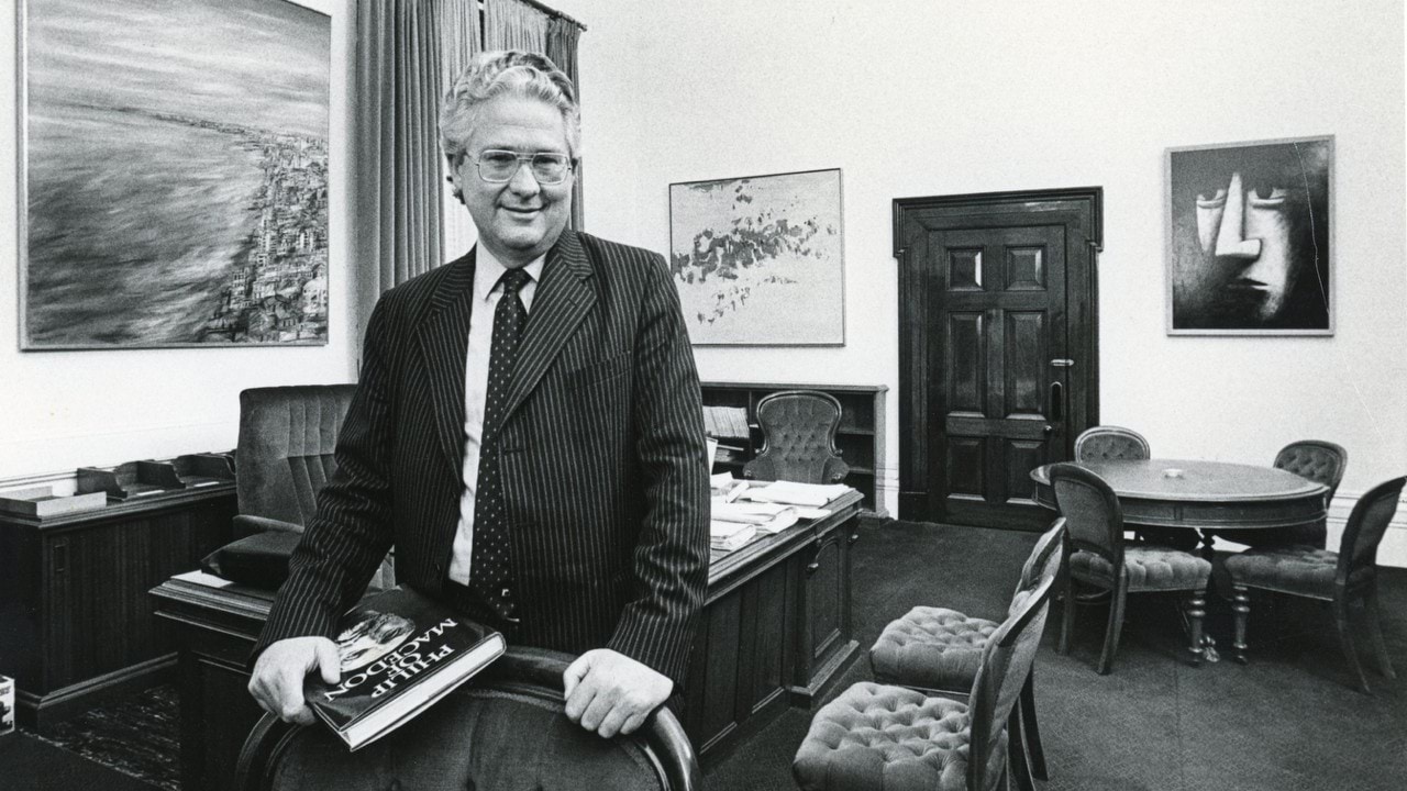 The Honourable Dr Race Mathews is wearing a pin-stripe suit and glasses. He leans on the back of a chair in his office holding a book titled Philip of Macedon