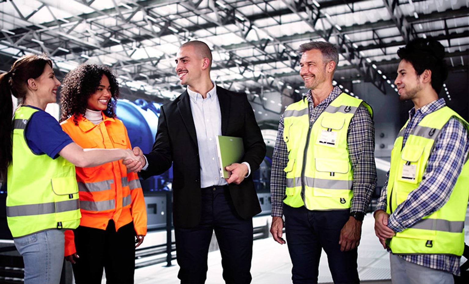 Five people in a manufacturing facility. Four are wearing high vis vests, one is wearing a suit. 