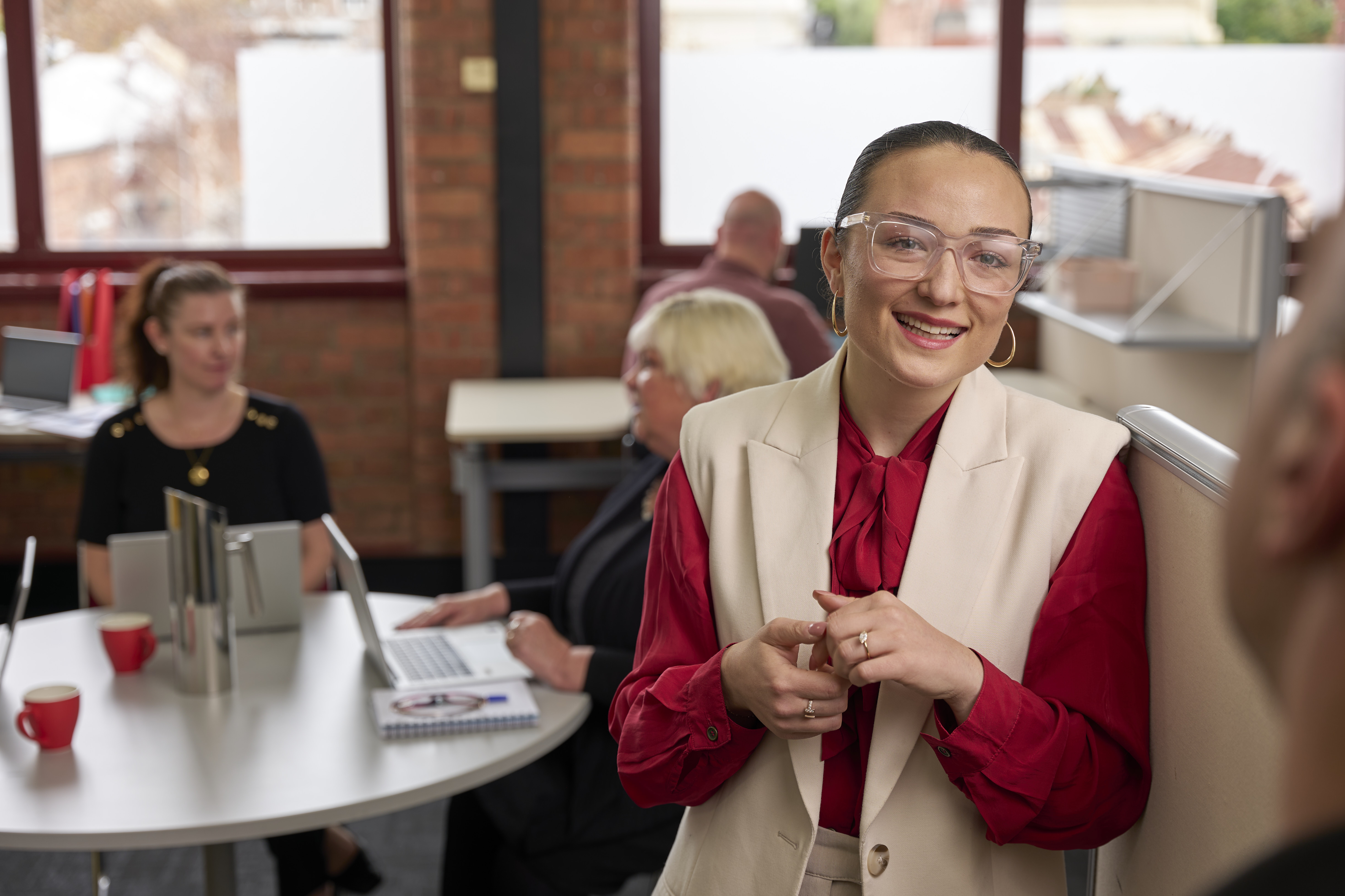 A person smiles while having a conversation with some one outside of the frame. there are people in the background in an office environment working.