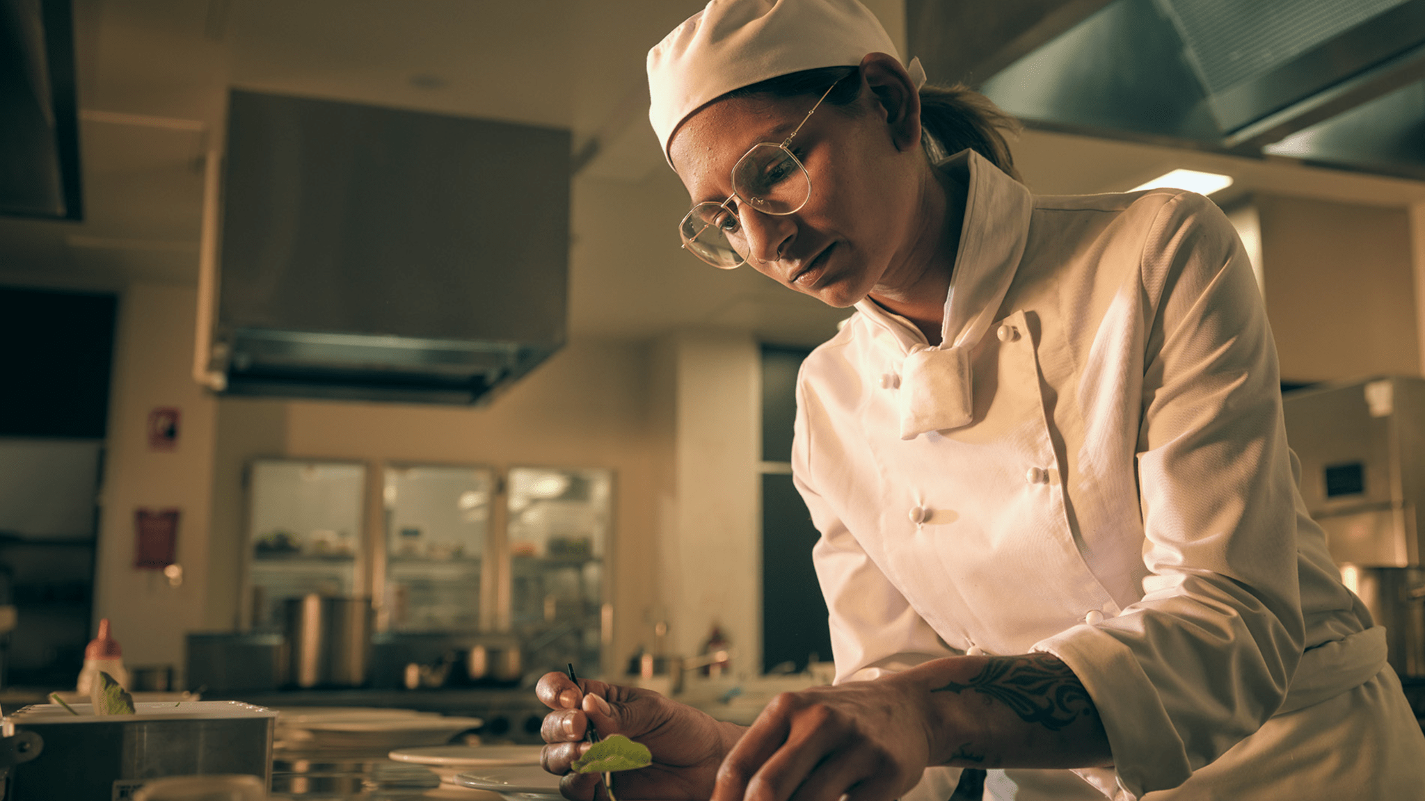A person in a chef's uniform uses tweezers to place food on a plate in a commercial kitchen.