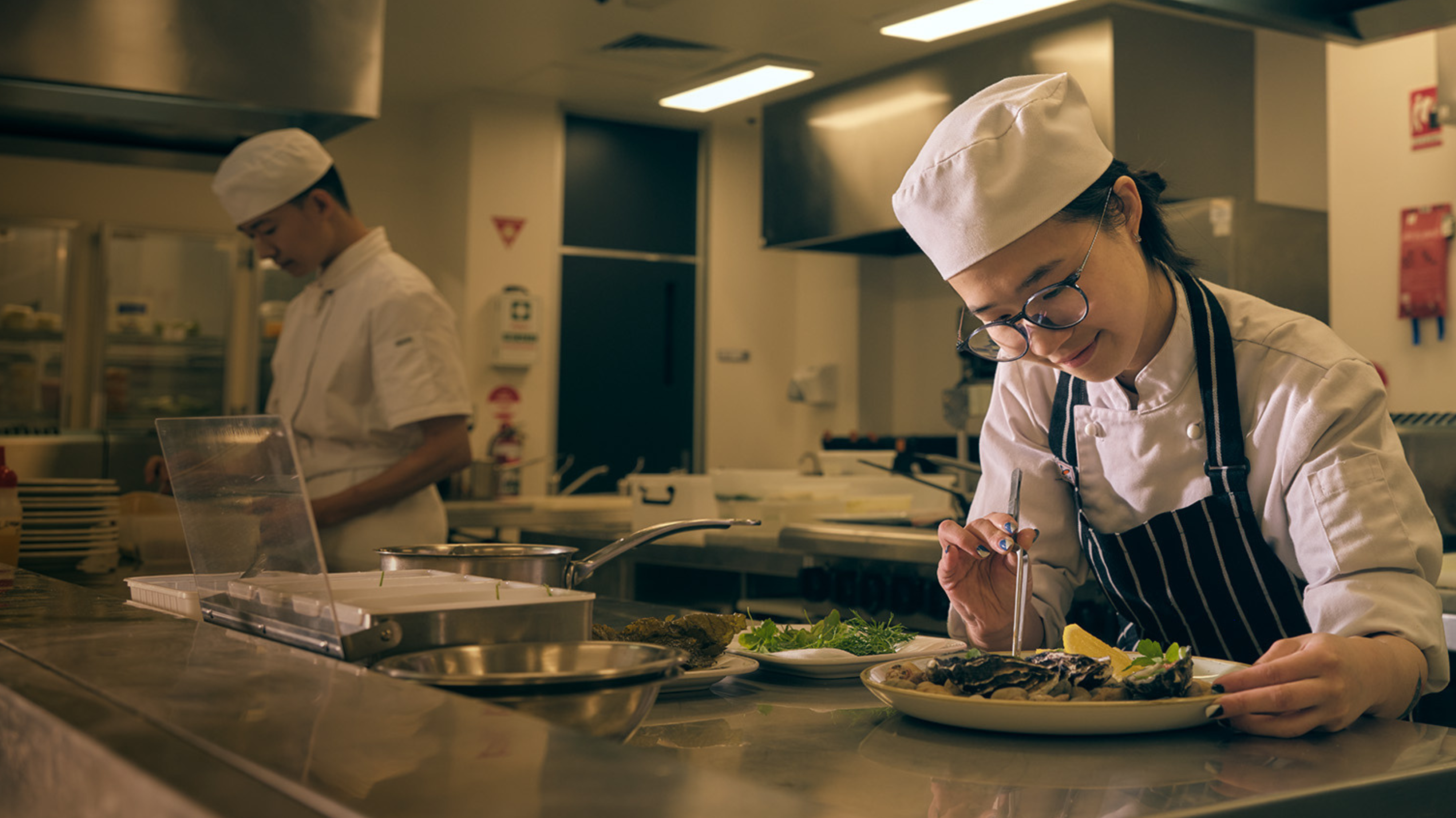A person in a chef's uniform and apron uses tweezers to place food on a plate in a commercial kitchen.