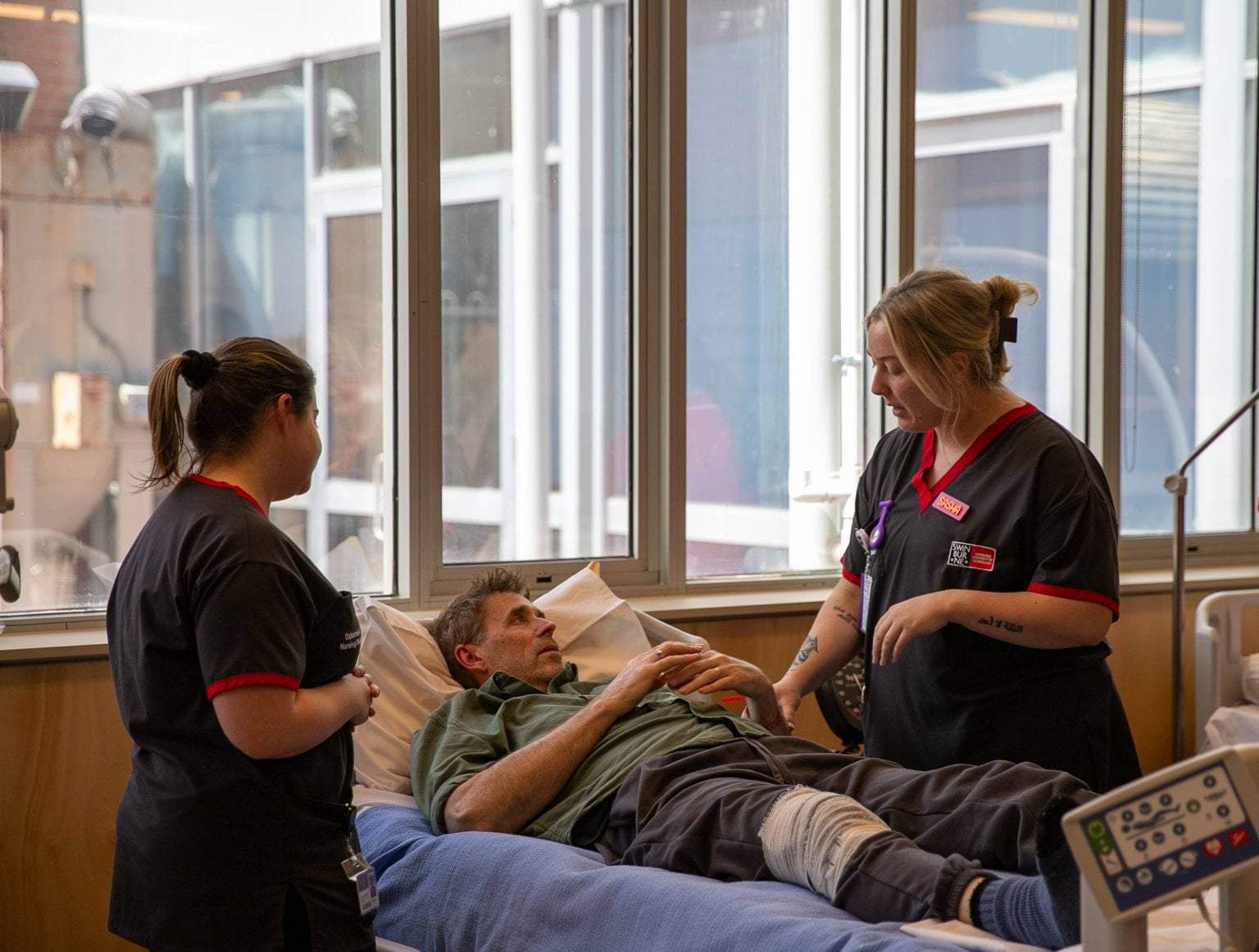 Student nurses dressed in scrubs talk to a patient in a hospital bed.