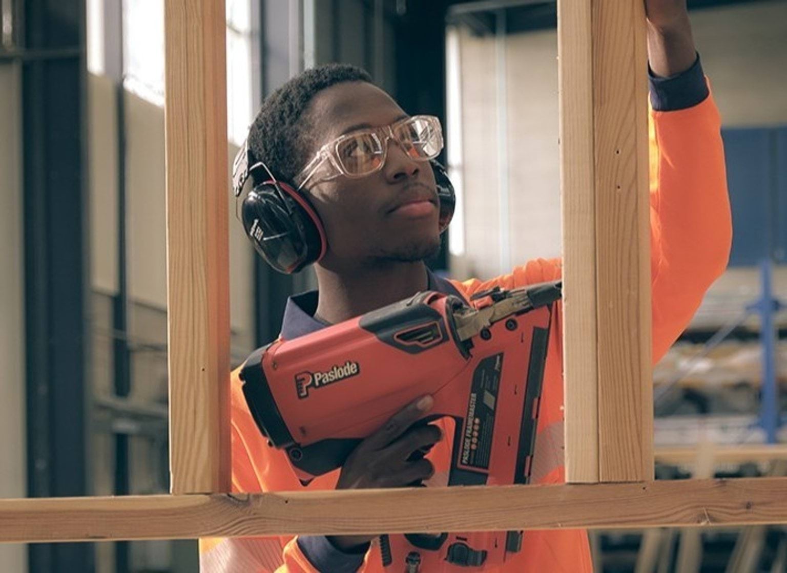 A person in hi-vis, headphones and safety glasses uses a nail gun on a wooden frame.