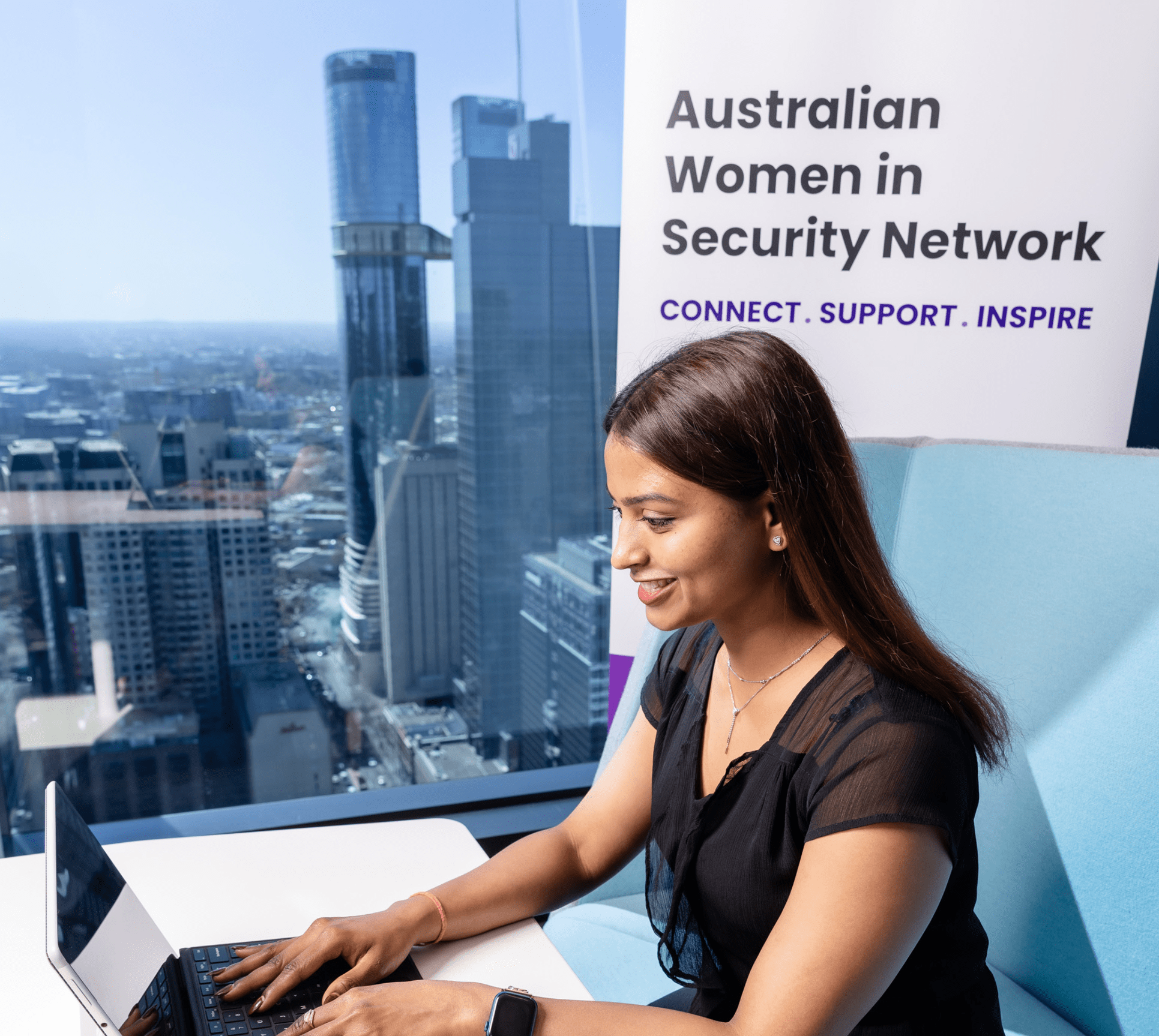 Padmini on a laptop in office with window to skyscrapers and a Australian Women in Security Network banner in the background