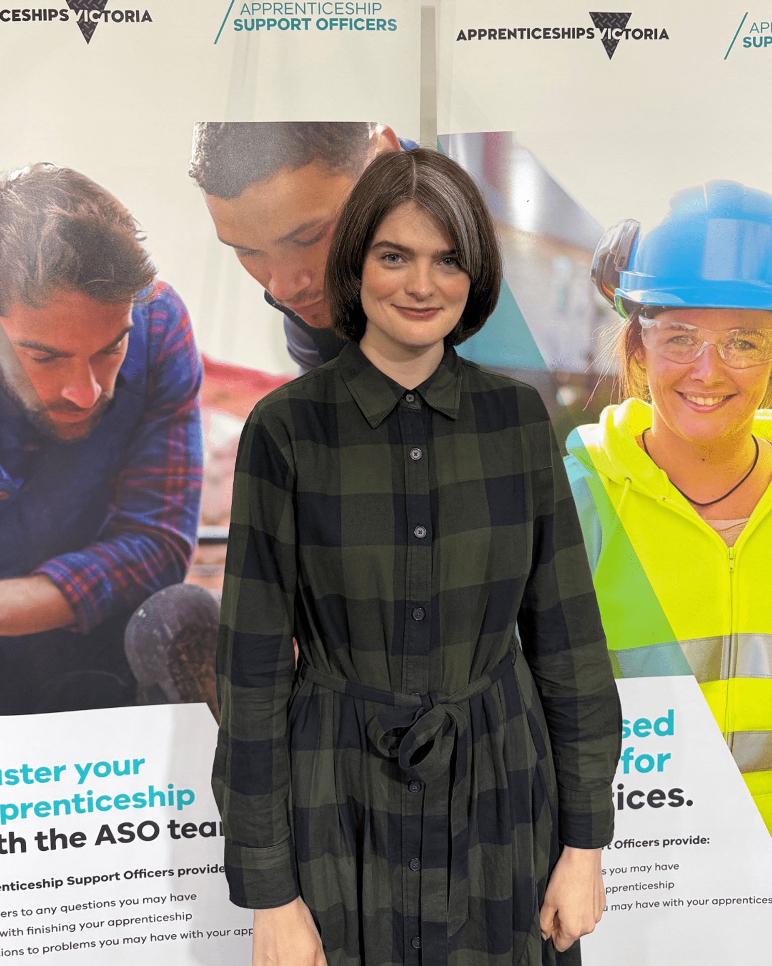 Alex is smiling in a checked dress, standing in front of an Apprenticeship Support Officer banner.