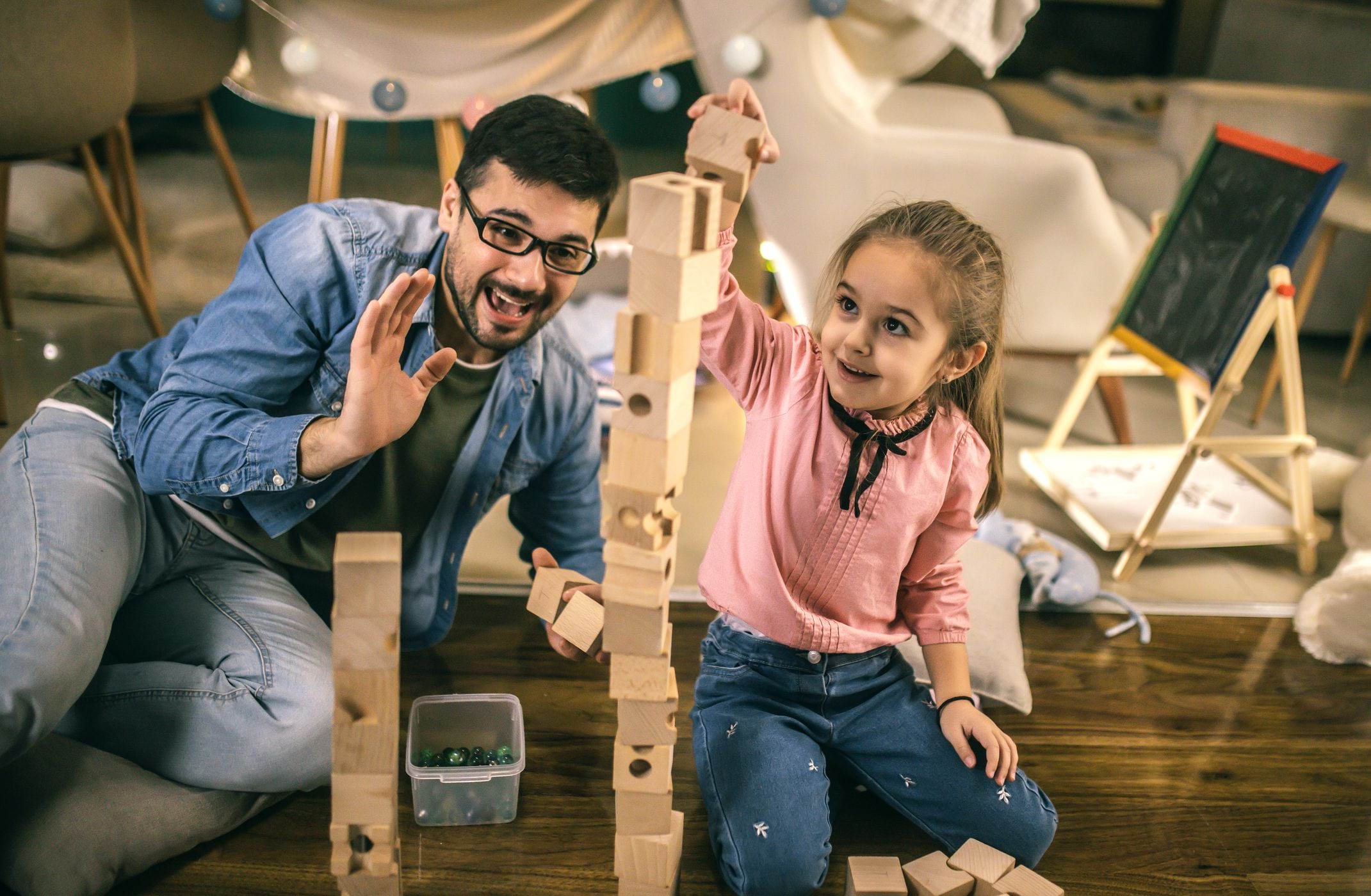 A man and a young girl build a tall wooden block tower on the floor, smiling in a living room.