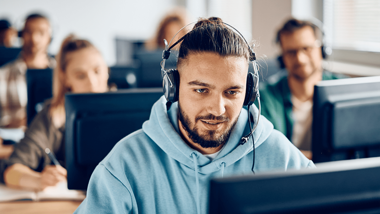 A person wearing headphones at a computer. Other people at computers are behind.
