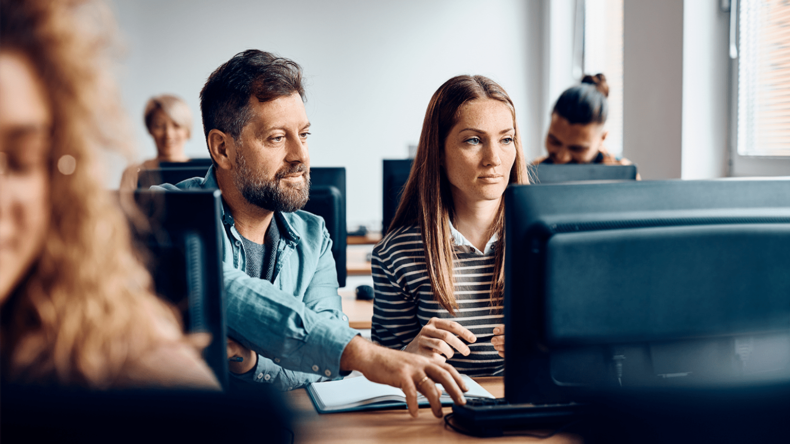 A person helping another person on a computer in a classroom of people on computers.