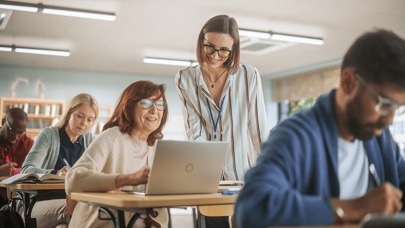 Students at desks in a classroom and a teacher standing and helping someone at a laptop.