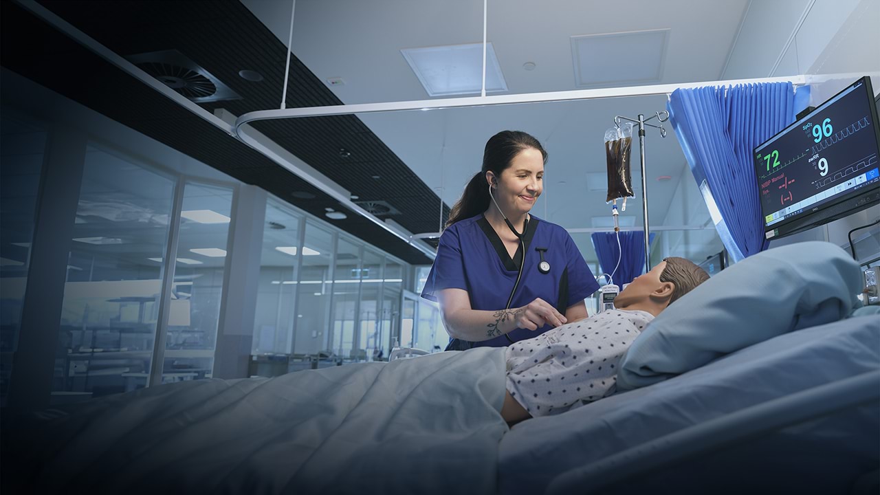 A person in a blue hospital uniform using a stethoscope on a medical mannequin in a hospital setting.