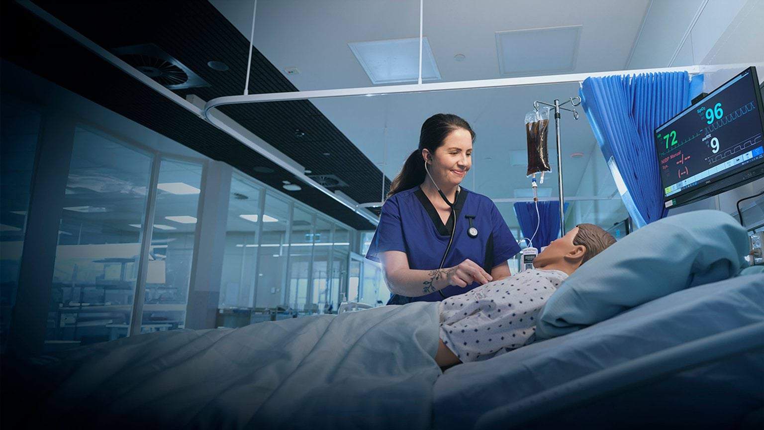 A person in a blue hospital uniform using a stethoscope on a medical mannequin in a hospital setting.