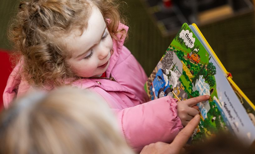 A child pointing on an illustration while being assisted by their educator to read a picture book